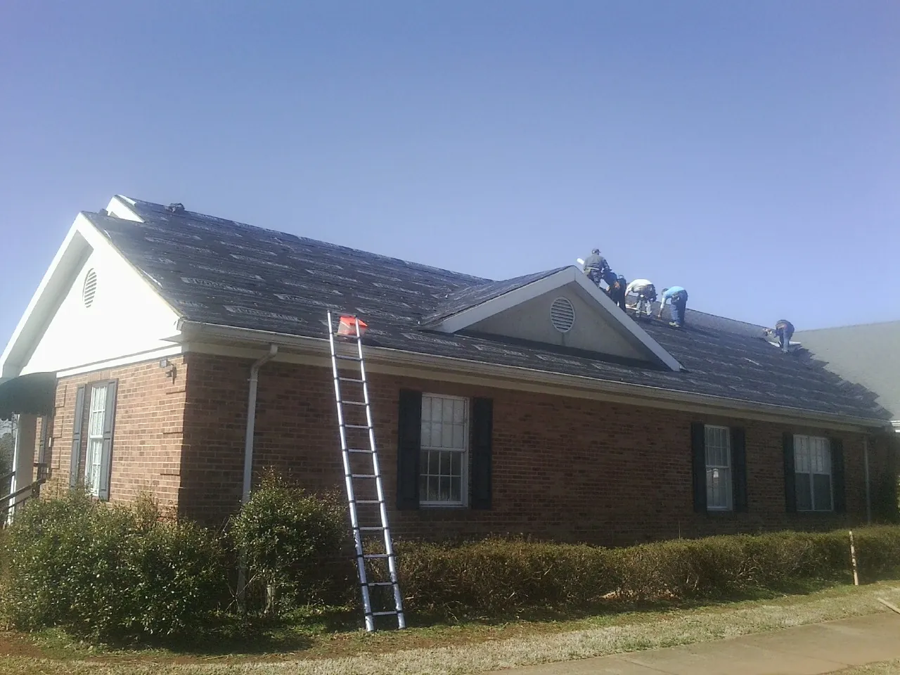 Crew tearing shingles off a brick ranch roof with a ladder set — Metal Roof Repair work by our crew in Clayton